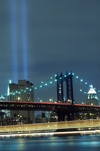 Boats pass under the Manhattan Bridge as they glide along the East River and lower Manhattan. The Tribute lights shine brightly...